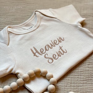 Puede incluir: Un body blanco para beb&eacute; con las palabras "Heaven Sent" bordadas en hilo marr&oacute;n. El body est&aacute; sobre una superficie de tela beige con un collar de cuentas de madera.
