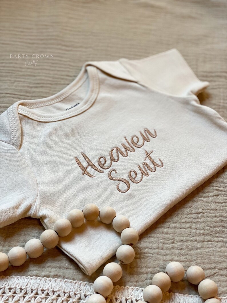 Puede incluir: Un body blanco para beb&eacute; con las palabras "Heaven Sent" bordadas en hilo marr&oacute;n. El body est&aacute; sobre una superficie de tela beige con un collar de cuentas de madera.