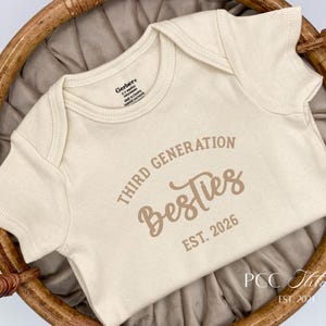 May include: A cream-colored baby onesie with the text "Third Generation Besties EST. 2026" in brown lettering. The onesie is made of soft material and is displayed in a woven basket.