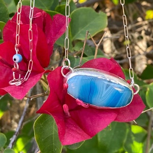 Pode incluir: Um colar de corrente de prata com um pingente de ágata azul. O pingente tem formato oval e tem uma moldura de prata. O colar está pendurado em um galho com flores rosa.