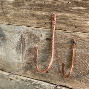 May include: Two hammered copper hooks mounted on a rustic wooden wall. The hooks are shaped like a 'J' and have a hammered texture.