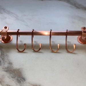May include: A copper kitchen utensil rack with five hooks. The rack is mounted to a wall with a round base.