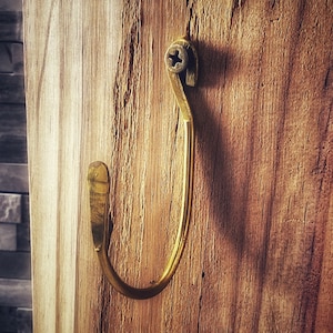 May include: A gold-colored metal hook mounted on a wooden surface. The hook is curved and has a simple design.