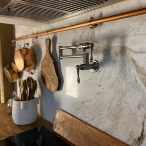 May include: A stainless steel kitchen tap with a pull-down spray mounted on a white marble backsplash. A copper pipe is mounted above the tap and wooden chopping boards and utensils hang from hooks.