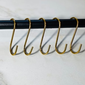 May include: Six gold-toned metal hooks hang from a black metal rod. The hooks are all the same size and shape, and they are arranged in a row.