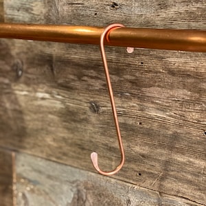 May include: A copper-colored S-shaped hook hangs from a copper pipe. The hook is attached to a wooden wall.