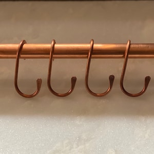 May include: Four copper hooks hang from a copper pipe. The hooks are all the same size and shape, and they are arranged in a row.