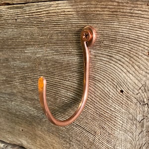 May include: A copper-colored metal hook with a screw attachment. The hook is mounted on a wooden surface.