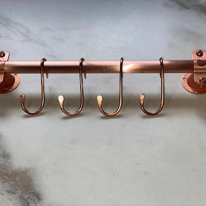 May include: Copper kitchen utensil rack with five hooks. The rack is mounted to a wall with two round brackets. The rack is made of copper pipe and has a polished finish.