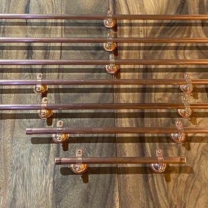 May include: A set of eight copper curtain poles of varying lengths, each with two decorative finials. The poles are arranged in a staggered pattern on a wooden surface.