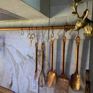 May include: A set of six copper kitchen utensils hanging from a copper pipe. The utensils include a whisk, a ladle, a slotted spoon, a spoon, and two spatulas. The utensils are all in a rustic style and have a hammered finish.