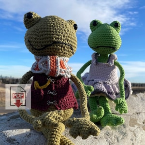 Peut inclure: Deux peluches de grenouilles au crochet assises sur un rocher. La grenouille de gauche porte un gilet bordeaux, une écharpe jaune et une écharpe blanche et orange. La grenouille de droite porte une robe lavande.