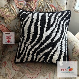 May include: Black and white zebra patterned crochet cushion on a floral patterned sofa. The cushion is square shaped and has a simple design. The text "TOY Recollection Crochet" is visible in the bottom right corner of the image.