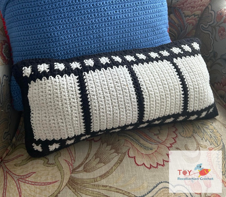 May include: A white and black crochet pillow with a film strip design. The pillow is sitting on a floral patterned couch.