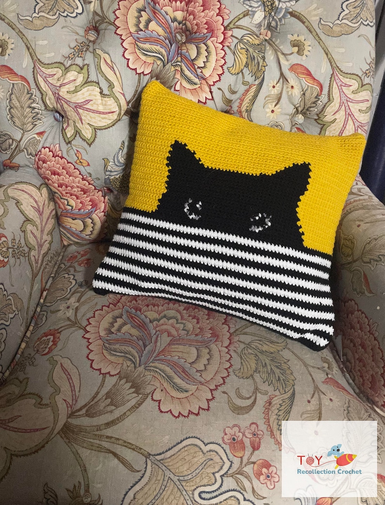 May include: A crocheted pillow with a black cat face on a yellow background and black and white stripes on the bottom half. The pillow is sitting on a floral patterned chair.