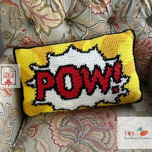 May include: A yellow, red and black crocheted cushion with the word "POW!" in a comic book style font. The cushion is sitting on a floral patterned armchair.