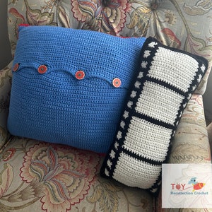 May include: Two crocheted pillows, one blue with red buttons and one black and white with a film strip design. The pillows are on a floral patterned couch.