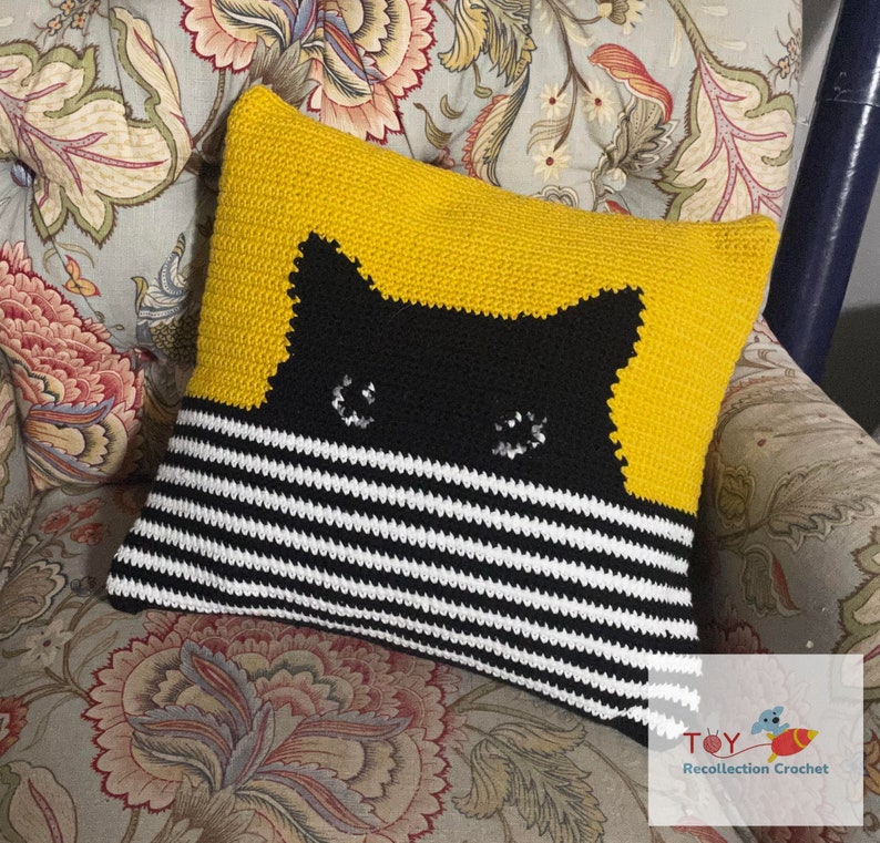 May include: A yellow and black striped pillow with a black cat face peeking out from behind the stripes. The cat has white eyes and a black nose.