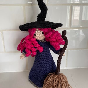 May include: A crocheted witch doll with pink hair, a black hat, and a blue dress. The doll is holding a brown broom.
