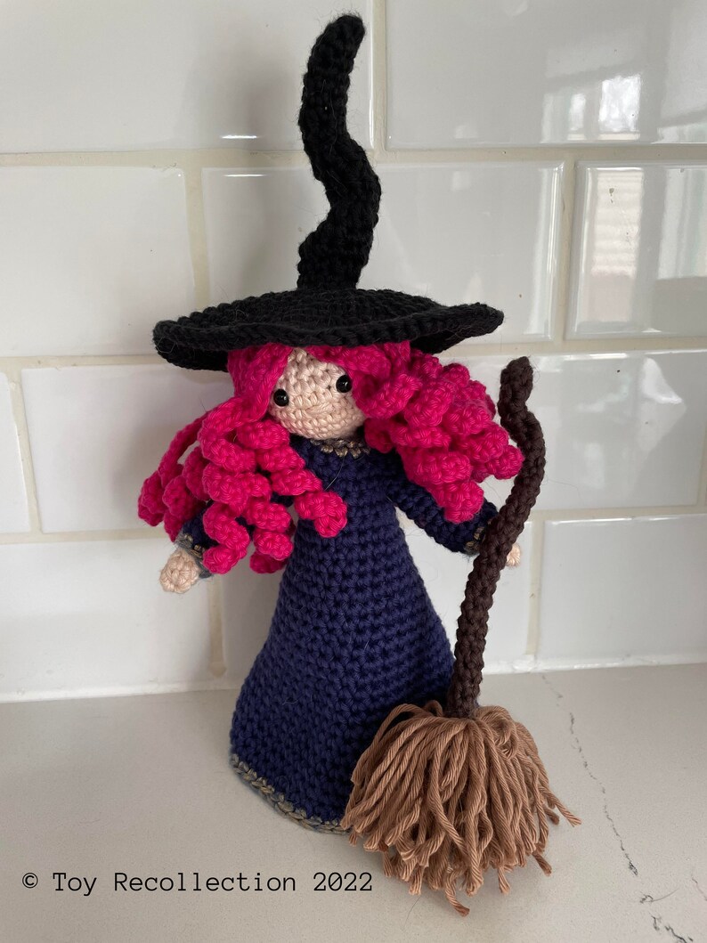 May include: A crocheted witch doll with pink hair, a black hat, and a blue dress. The doll is holding a brown broom.