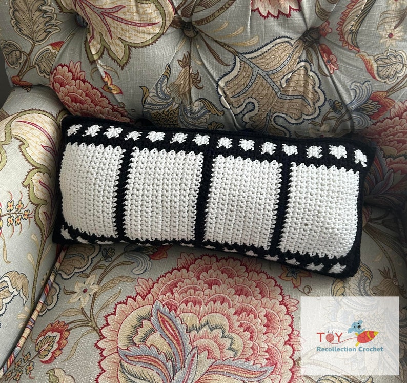 May include: A black and white crocheted pillow with a film strip design. The pillow is sitting on a floral patterned armchair.
