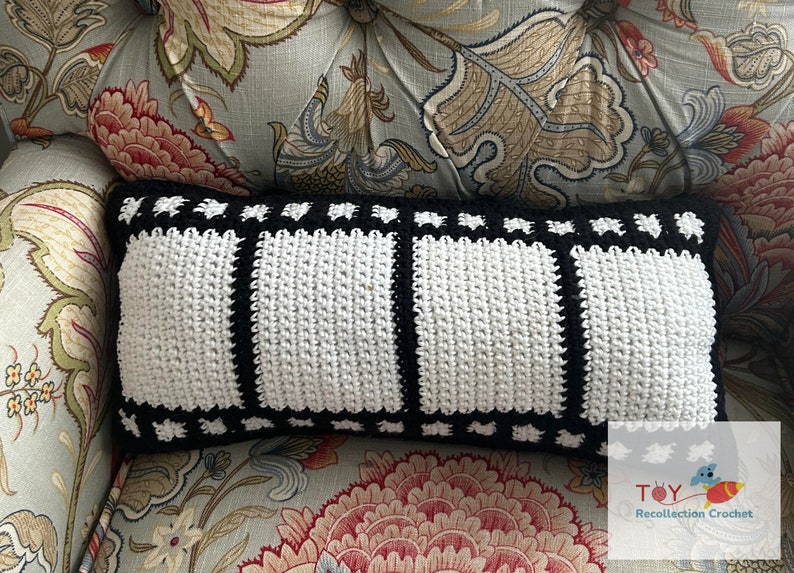 May include: A black and white crocheted pillow with a film strip design. The pillow is sitting on a floral patterned couch.