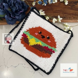 May include: Two white crocheted pot holders with black borders, featuring a cartoon burger with a smiling face. The pot holders are made with different colored yarn to create the details of the burger, including a red tomato, green lettuce, and yellow cheese. The text "Toy Recollection Crochet" is visible in the bottom right corner of the image.