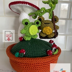 May include: Crocheted amigurumi toy set featuring two green frogs on a swing, a mushroom with a red and white cap, and a green base with red berries. The base is in a brown pot. The set is labeled "Toy Recollection Crochet" and includes a PDF download icon.