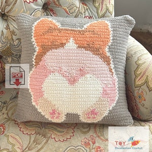 May include: A crocheted pillow with a gray background and a pink and orange hamster design. The pillow is sitting on a floral patterned couch.