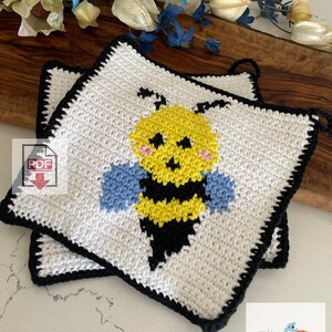 May include: Two crocheted white pot holders with black trim, featuring a yellow and black bee with blue wings. The pot holders are stacked on top of each other. The image includes the text "TOY Recollection Crochet".