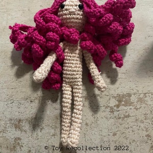 May include: A crocheted doll with long pink hair and a beige body. The doll has black eyes and a simple face.