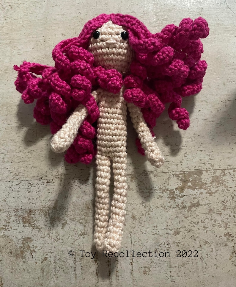 May include: A crocheted doll with long pink hair and a beige body. The doll has black eyes and a simple face.