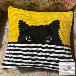 May include: A yellow and black crocheted pillow with a black cat face design. The cat's eyes are white and the pillow has black and white stripes on the bottom half.
