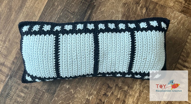 May include: A long, rectangular pillow with a black and white film strip design. The pillow is made of crocheted yarn and has a light blue center.