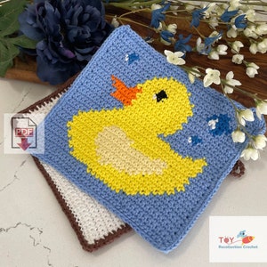 May include: Two crocheted pot holders, one blue with a yellow duck and the other white with a brown border. The blue pot holder has a pattern of small blue fish. The pot holders are on a wooden surface with a flower and a sign that says "Toy Recollection Crochet".
