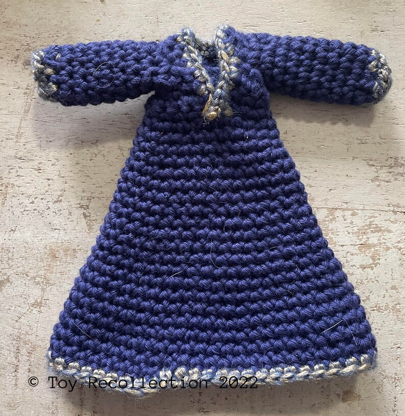 May include: A crocheted doll's dress in a deep blue colour with a white and gold trim. The dress has long sleeves and a V-neck.