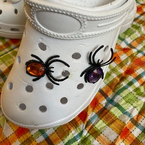 May include: White clogs with black spider embellishments and jeweled accents. The spiders have orange and purple gems for eyes.