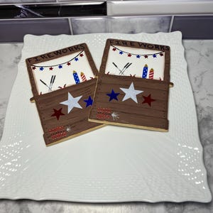 May include: Two wooden, brown-colored boxes with the word "FIREWORKS" on top, decorated with red, white, and blue stars and fireworks imagery. The boxes are placed on a white, textured, square plate.