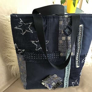 Peut inclure: Un sac cabas bleu marine avec des détails en patchwork et une bandoulière noire. Le sac est fait de différents tissus avec une variété de textures et de motifs, y compris des étoiles, des rayures et du tartan.