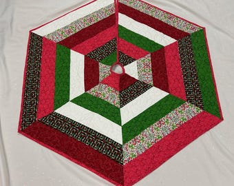 May include: A quilted Christmas tree skirt in a hexagonal shape. The design features alternating sections of red, white, and green fabric, with accents of patterned fabric. The skirt has a circular opening in the center.