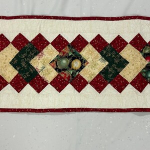 May include: A quilted Christmas table runner with a cream-colored center and red diamond-shaped accents. The center features alternating squares of green, tan, and red fabric with festive fruit and foliage patterns. The runner is approximately 14 inches wide and 40 inches long.