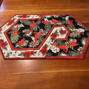 May include: A decorative table runner with a hexagonal shape. The runner features a black background with red cardinals, pinecones, and poinsettias. The edges are trimmed with a red border, and the overall design has a festive, seasonal theme.