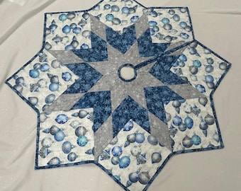 May include: A star-shaped quilted tree skirt with a central hole. The design features a gray star in the center, surrounded by blue and white patterned sections with snowflake and ornament motifs. The edges are finished with a dark blue binding.