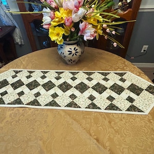 May include: A white and green quilted table runner with a diamond pattern. The runner is on a gold table with a vase of pink and yellow flowers.