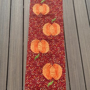 May include: A rectangular, quilted table runner featuring four orange pumpkins with green leaves on a burgundy background. The pumpkins are the focal point, with detailed stitching. The background has a floral pattern.