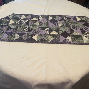 May include: A purple and green quilted table runner with a geometric pattern. The runner is made of fabric squares sewn together to create a patchwork design.