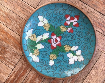 May include: A round, blue and white porcelain plate with a floral design. The plate features a repeating pattern of stylised flowers and leaves in shades of red, white, green, and yellow.