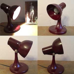 May include: A vintage desk lamp with a maroon-coloured base and shade. The lamp has a flexible arm and a light bulb. The lamp is on a wooden surface.