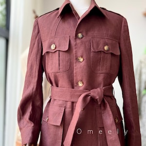 Puede incluir: Chaqueta de lino color granate con cuatro bolsillos delanteros, cinturón y botones. La chaqueta se exhibe en un maniquí y tiene un estilo safari clásico. El texto "Omeely" es visible en la chaqueta.