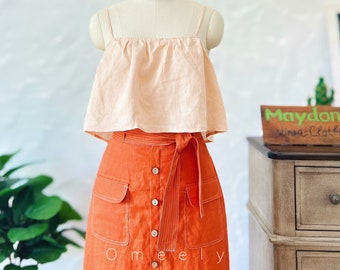 May include: A light peach-colored spaghetti strap crop top paired with a high-waisted, vibrant orange skirt. The skirt features a front button closure, two large patch pockets, and a matching fabric tie belt with white stitching details.